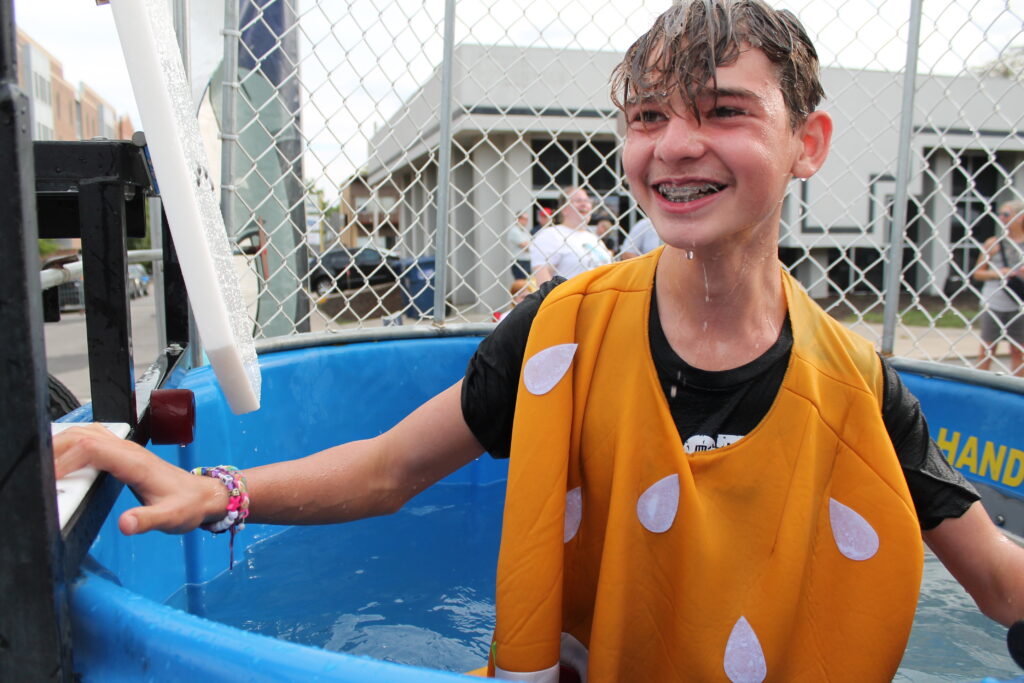 A young person in a yellow smock with white dots grins from a blue dunk tank where they have been dunked.