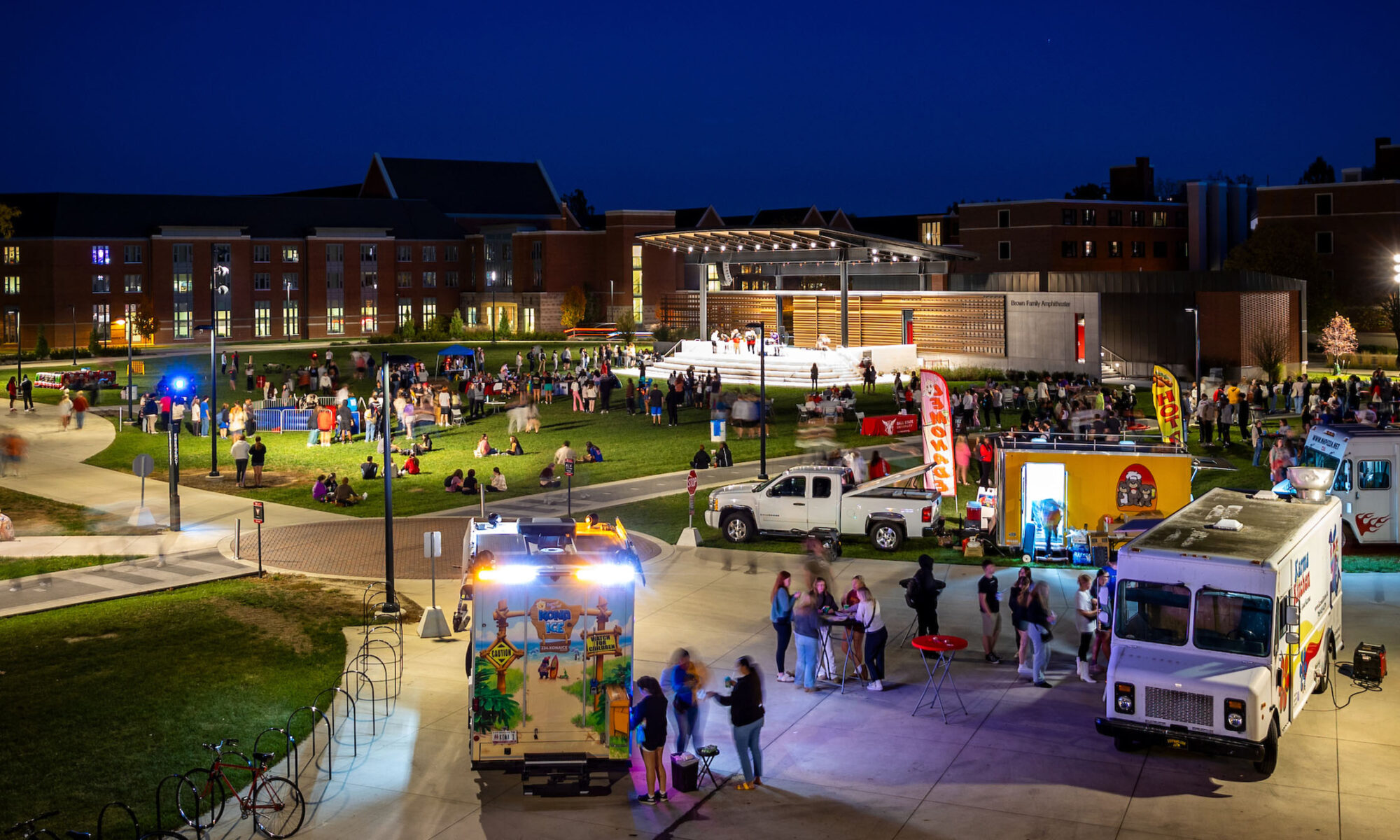 People gather at food trucks in the foreground with the amphitheater lit up in the background. the night sky is a deep blue and the grounds are brightly lit.