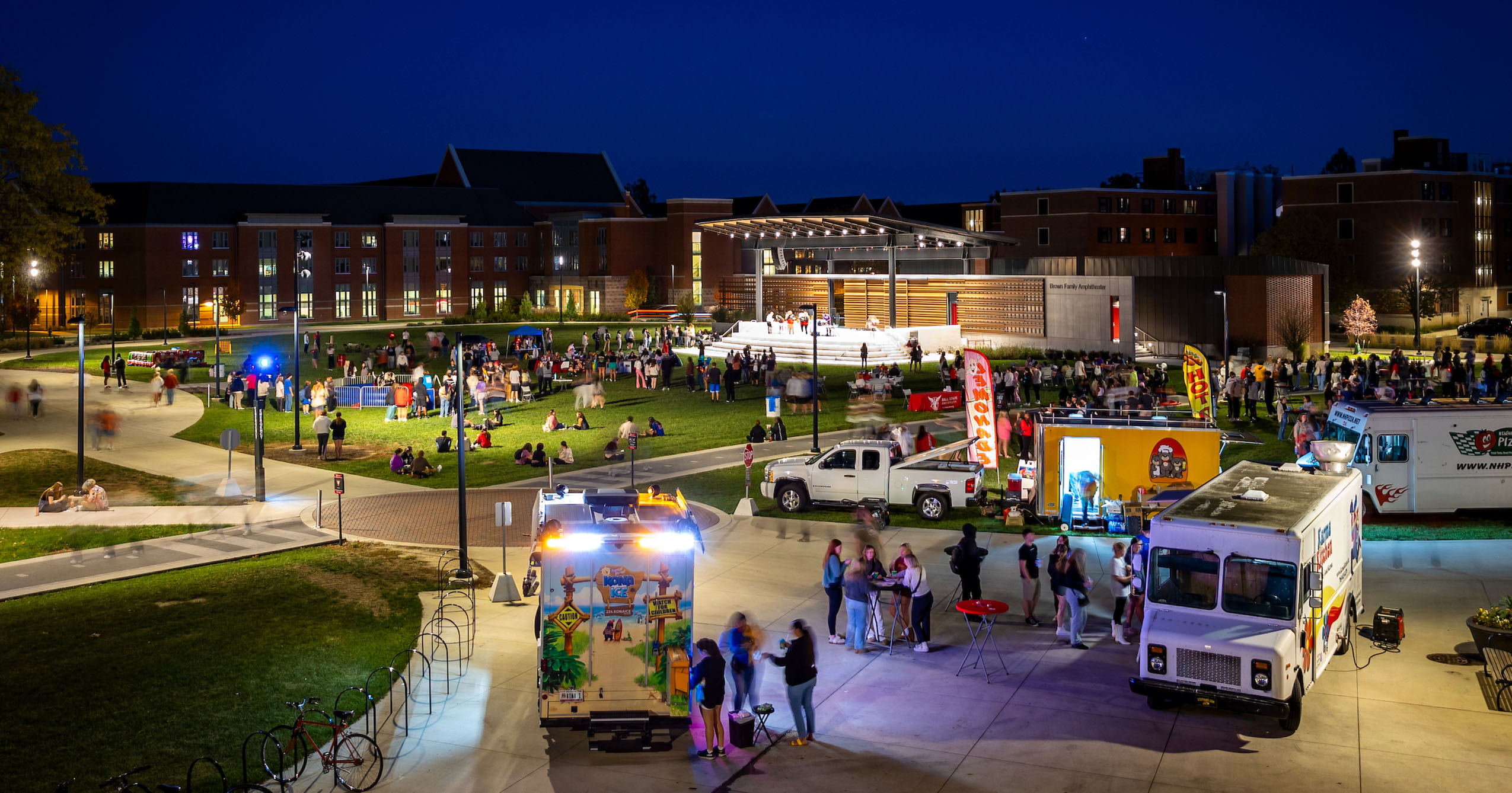 People gather at food trucks in the foreground with the amphitheater lit up in the background. the night sky is a deep blue and the grounds are brightly lit.