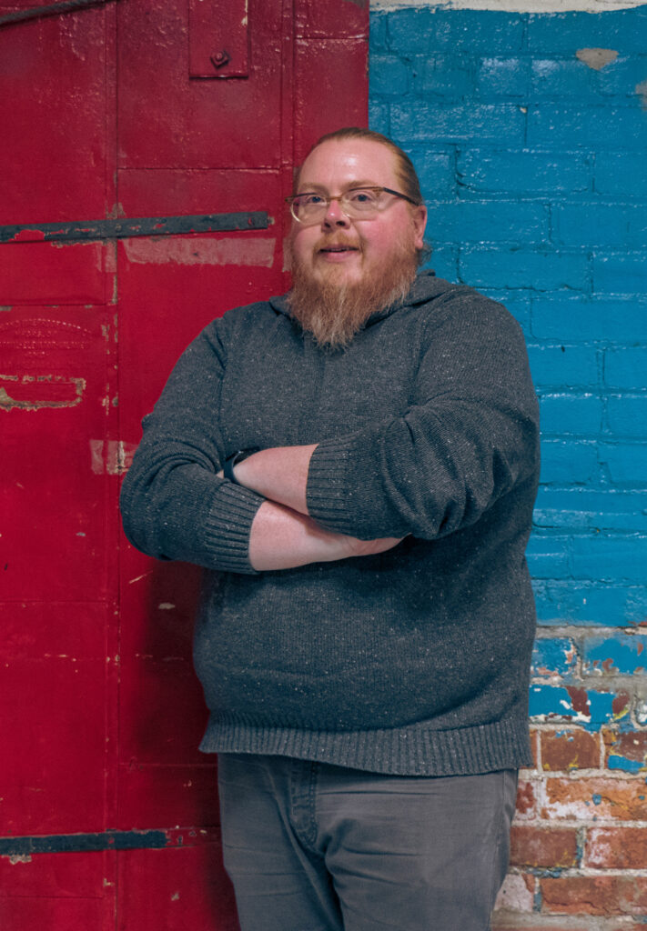 Lucas Pint, a man with a beard and crossed arms, leans confidently against a brick wall