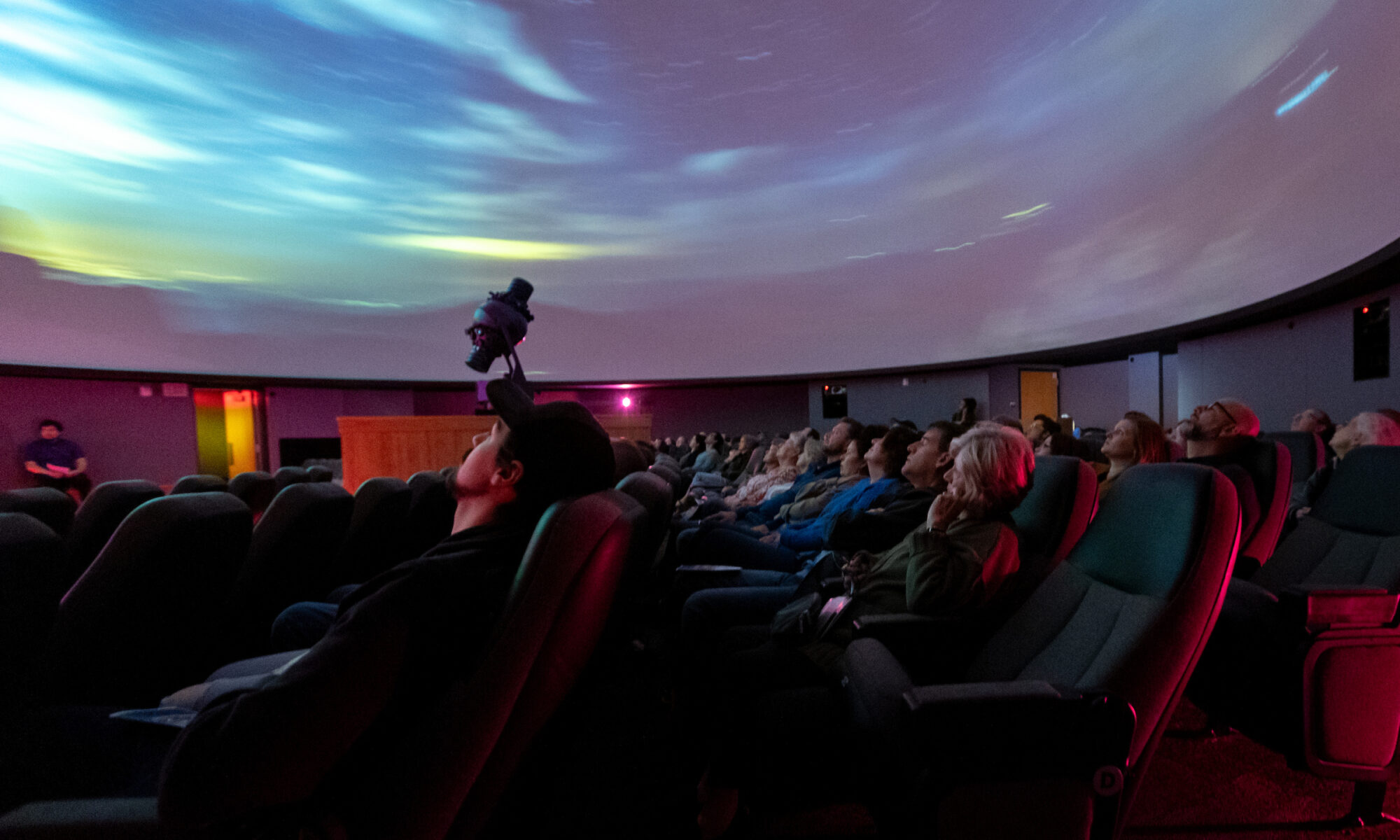 The audience at a planetarium show sit in seats tilted back to make it easier to see the bright swirls of colors displayed on the ceiling.