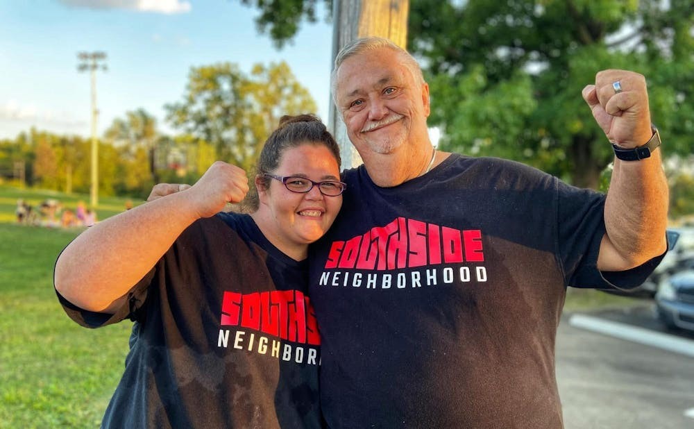 Two smiling people with their arms around each other stand in a sunny green space. They are both flexing and wearing Southside Neighborhood t-shirts