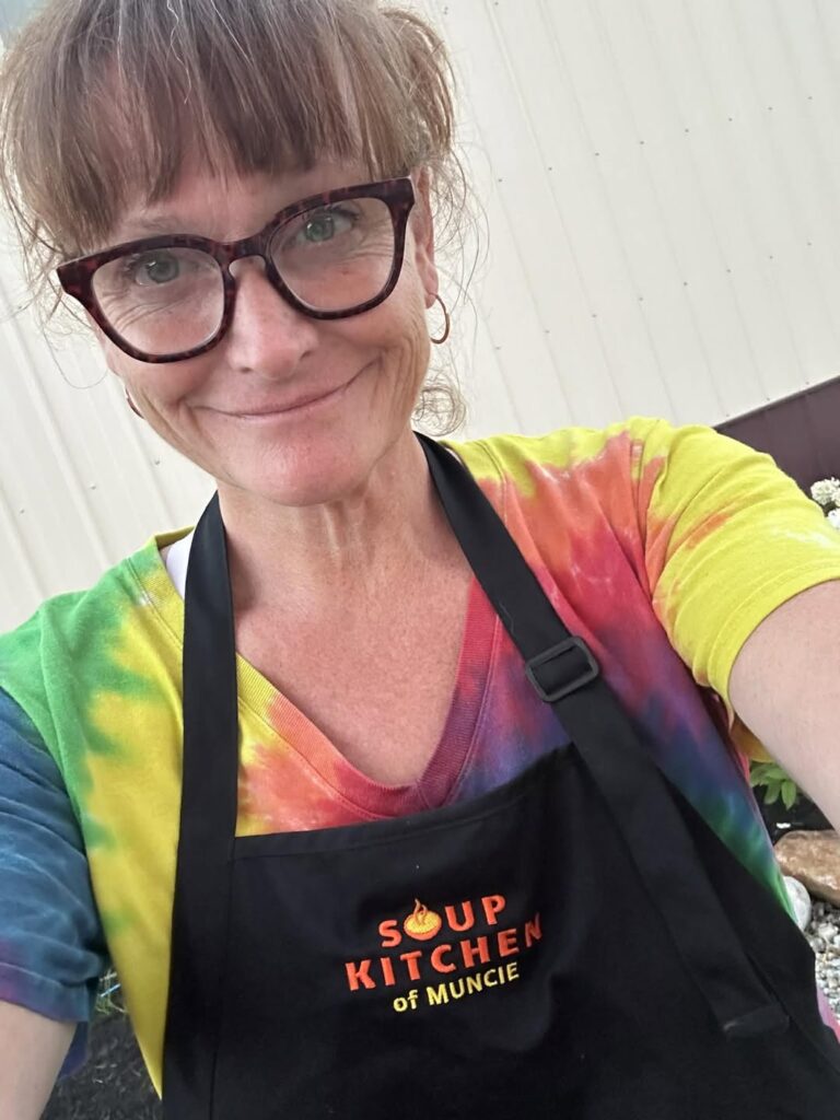 Christiana Mann selfie - she is wearing a rainbow tie dye shirt under a black apron with a "Soup Kitchen of Muncie" logo on it.
