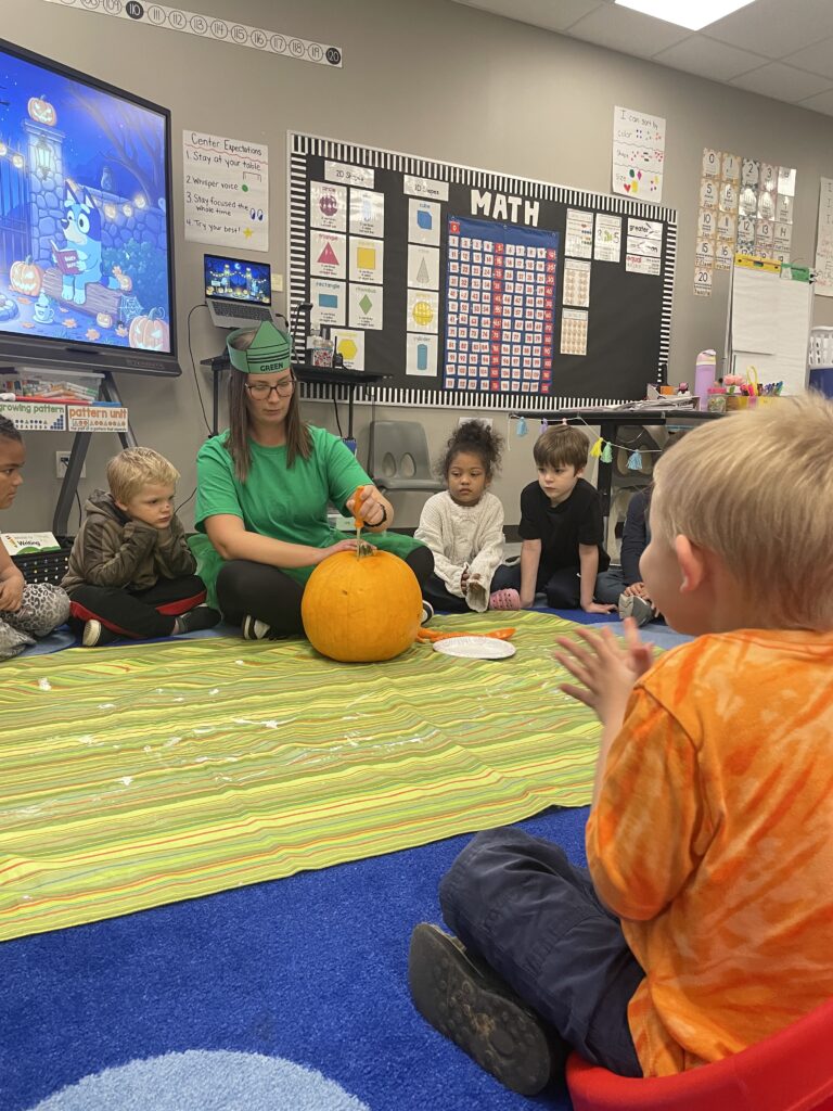 Mary Kult sits on a colorful floor surrounded by attentive children who watch her carve a pumpkin