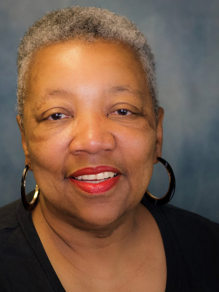 Headshot of Yvonne Thompson, a black woman with a bright smile wearing a black shirt and black and gold hoop earrings on a gray/blue background