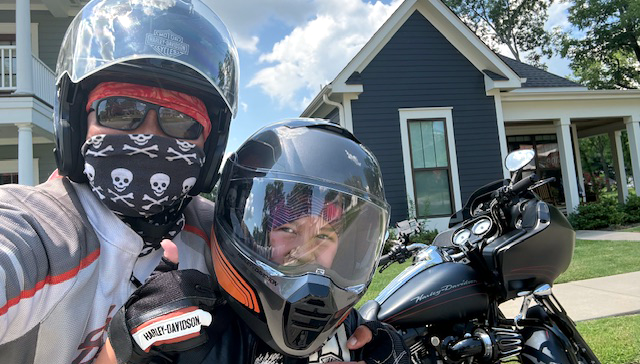 A woman and a child taking a selfie on a motorcycle in front of some houses. Both are smiling and wearing helmets.