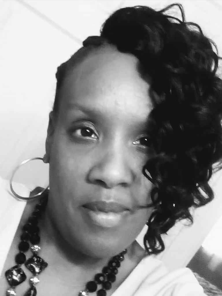 A black and white headshot of Trae Greenleaf, a black woman with a slight smile in a white blouse complimented by black jewelry.