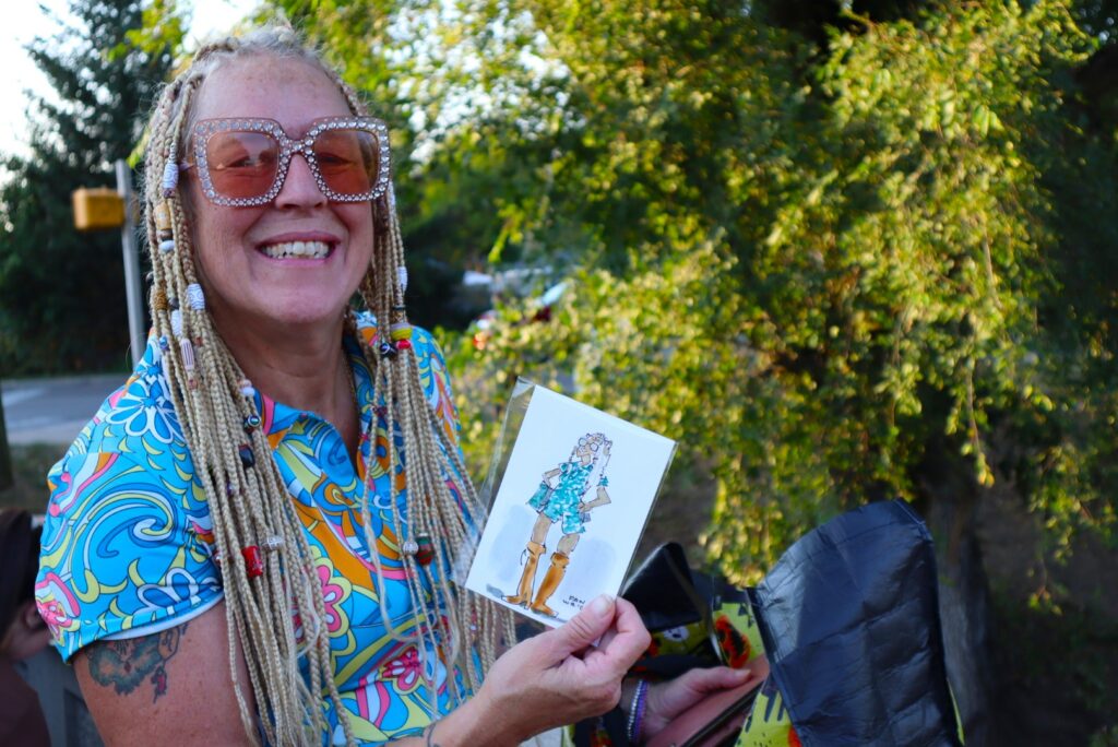 A smiling woman with long, beaded blond braids shows a card with a caricature of her on it to the camera.
