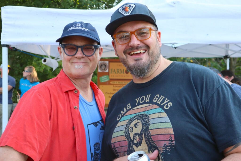 A woman in a red button down shirt over a light blue t-shirt and a man in a dark gray graphic t-shirt lean into each other and smile at the camera in front of a white vendor tent.
