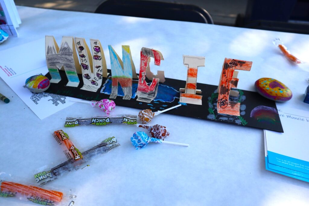 A colorful craft project of the cut out letters spelling "Muncie", each of them decorated with a different pattern in crayon. The stand up vertically off of a black paper base which has also been colored.