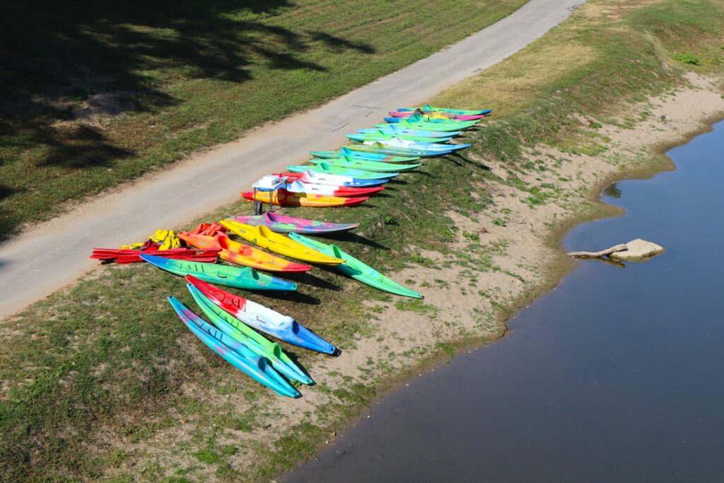 A photo taken from the bridge looking down on colorful kayaks piled up on the shore of the river between the trail and the water.