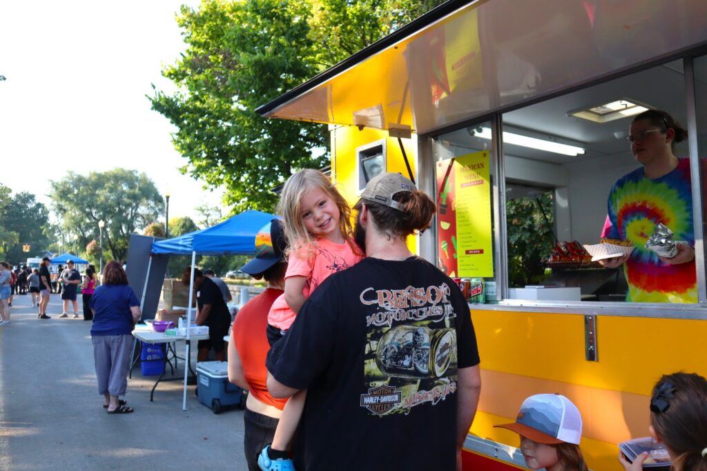 A small child grins over the shoulder of the adult holding her as they wait for food at a bright yellow food truck. The vendor at the window is wearing a rainbow tie-dye shirt and speaking with another customer.