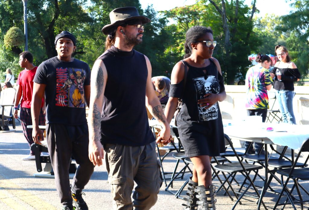 Three young mean in black graphic t-shirts walk together past a row of folding chairs on the bridge.