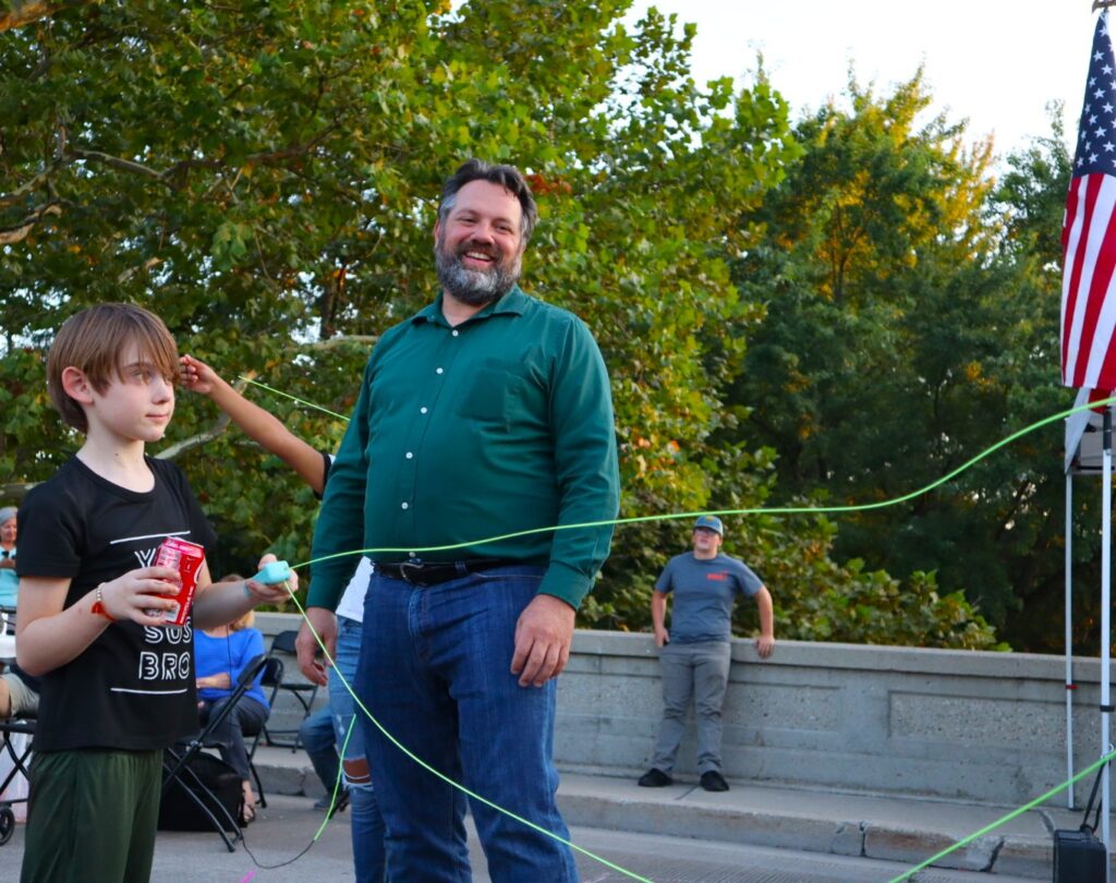A man in a green shirt laughs at the antics of a boy in a black shirt holding a can of soda and one end of a bright green loop of cord.