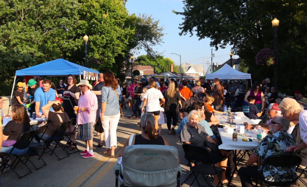 The bridge is packed with tents and vendors lining each edge, some with blue tents over their tables. Long tables are set up down the middle of the road with white paper covering the surfaces. Nearly every seat is filled with people eating and talking.