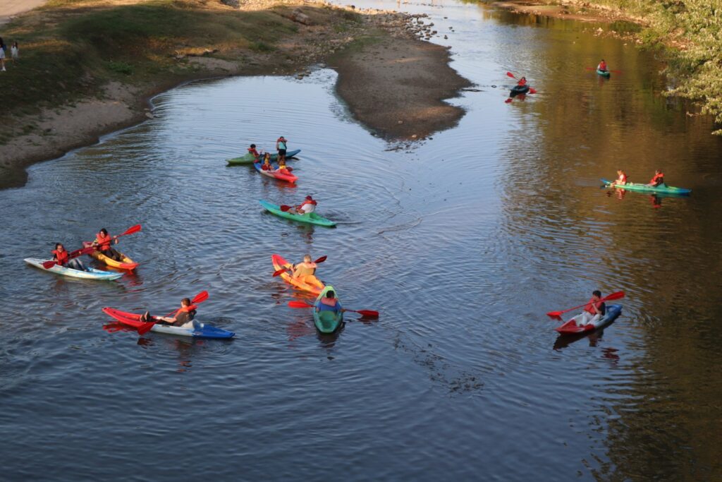 View of the river from the bridge. There is a hooked sandbar in the upper left. A group of people kayak in colorful boats in the water.