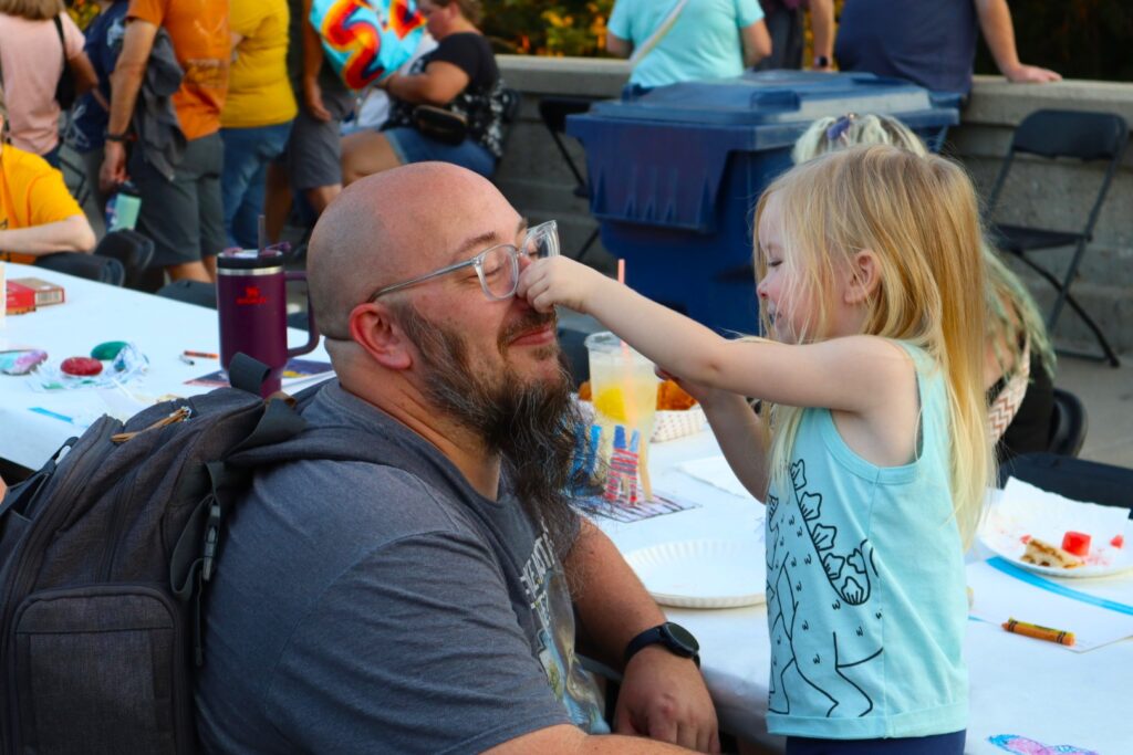 A small blond child in a teal shirt and matching hairbow grabs a bearded man's nose. The surface of one of the paper-covered tables set up down the center of the bridge is visible behind them.
