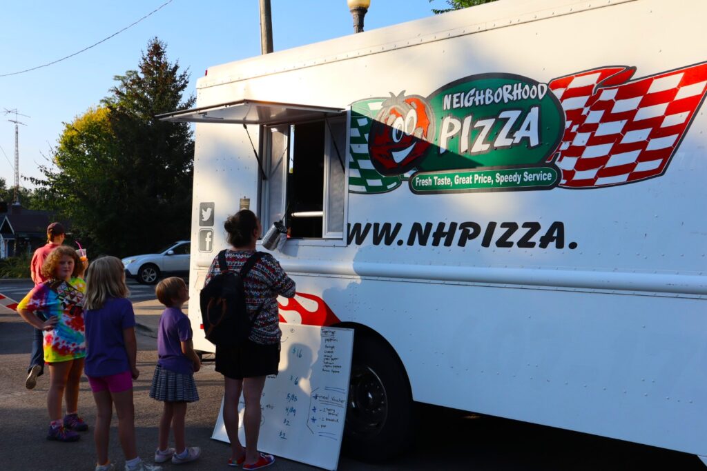 A group of people in shorts and t-shirts line up at a food truck selling pizza.