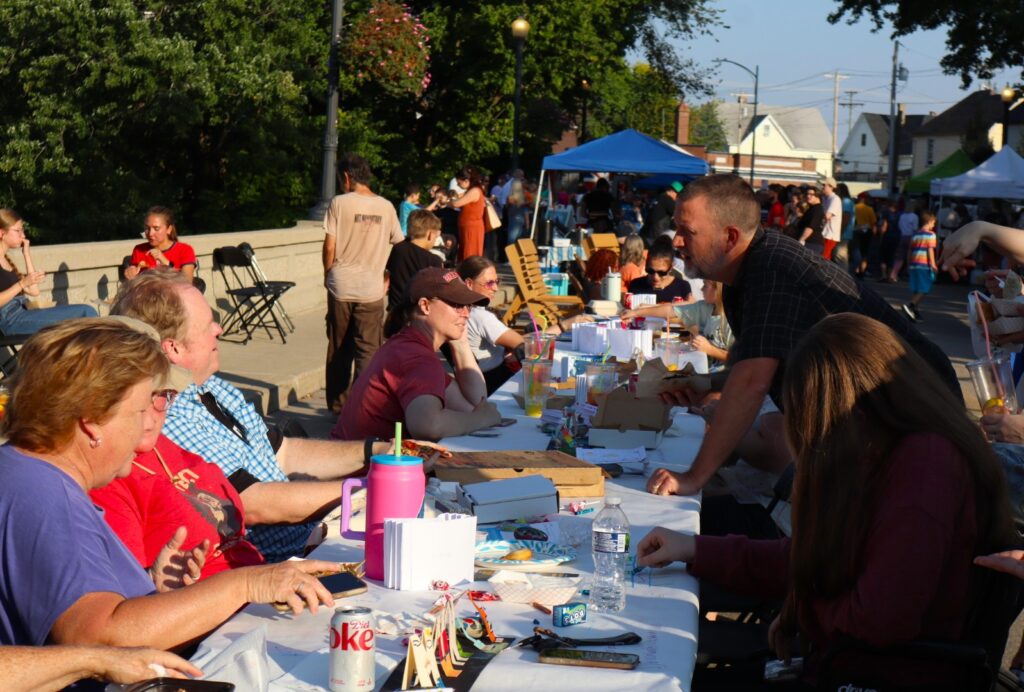 A group of people sit talking at one of the tables. The bridge barrier is visible in the background, and farther down, a blue tent covers a vendor table. 