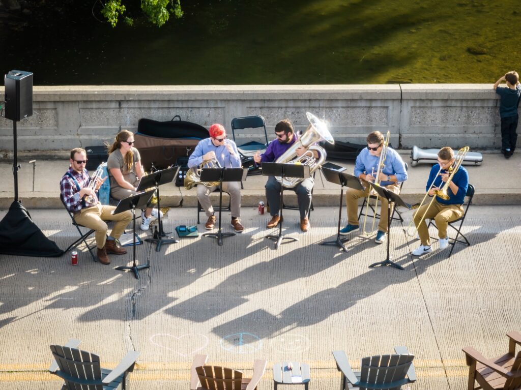 An aerial shot of a band setting up in a semi-circle at the edge of the road. The river is visible behind them, and an arc of lawn chairs is just visible at the bottom of the frame.