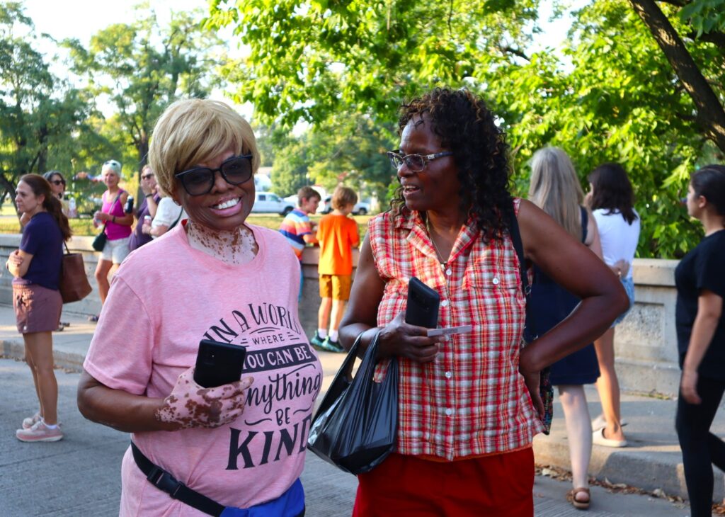 A woman in a pink shirt and sunglasses smiles at the camera while another woman next to her in a red striped shirt grins at her with a hand on her hip. The bridge is busy behind them.