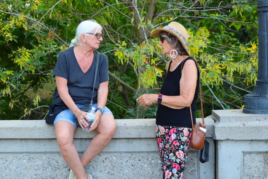 Two women speaking on the bridge. The one on the left is sitting on the barrier and the one on the right is turned toward her, leaning back on the barrier.