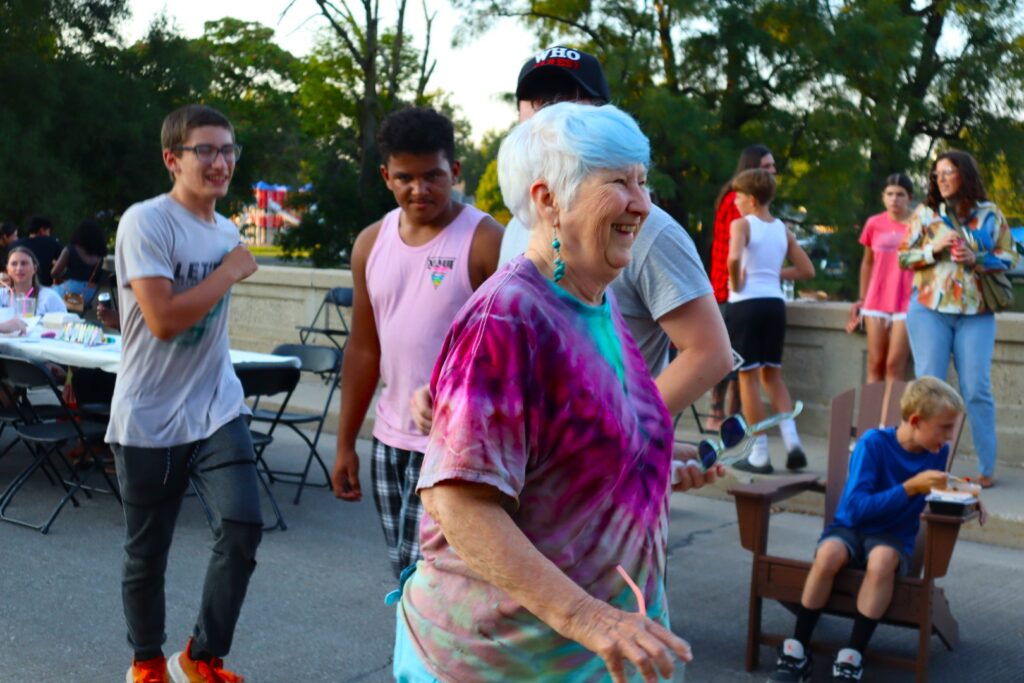 A woman with short, white hair walks down the bridge smiling. A variety of teenagers mingle in the background