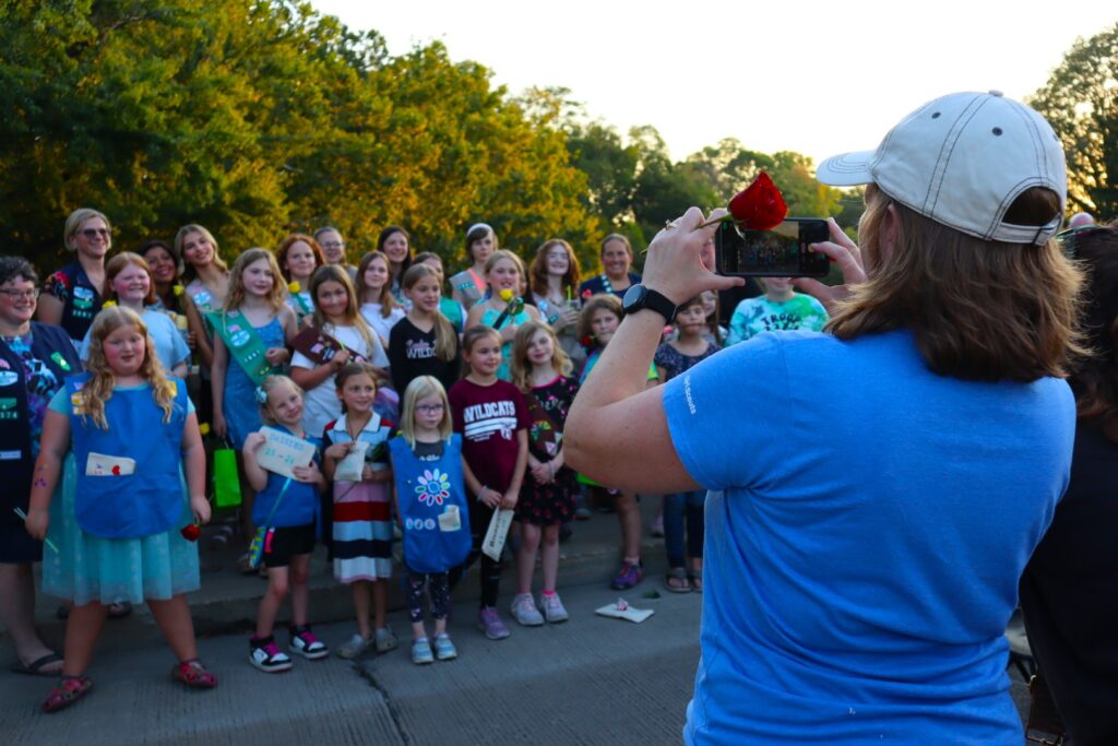 A woman in a blue shirt and white hat takes a group photo of children lined up against the bridge barrier. The trees of the river shoreline are behind them.