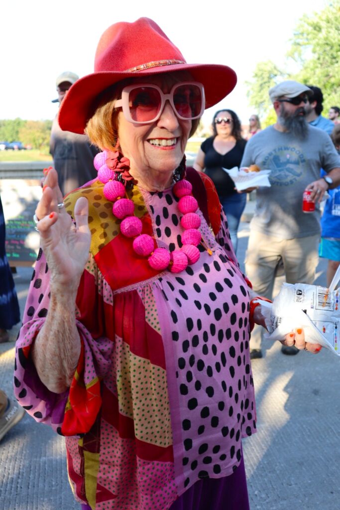 A smiling woman wearing pink from head to toe, including a bright pink hat, sunglasses, and a very large beaded necklace. She walks down the bridge with a crowd behind her.