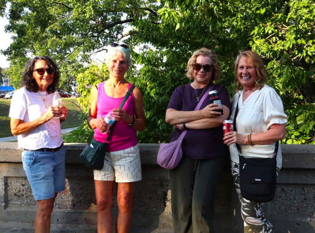 Four smiling women dressed for hot weather lean against the gray cement barrier lining the bridge. Bright green leaves make up the background