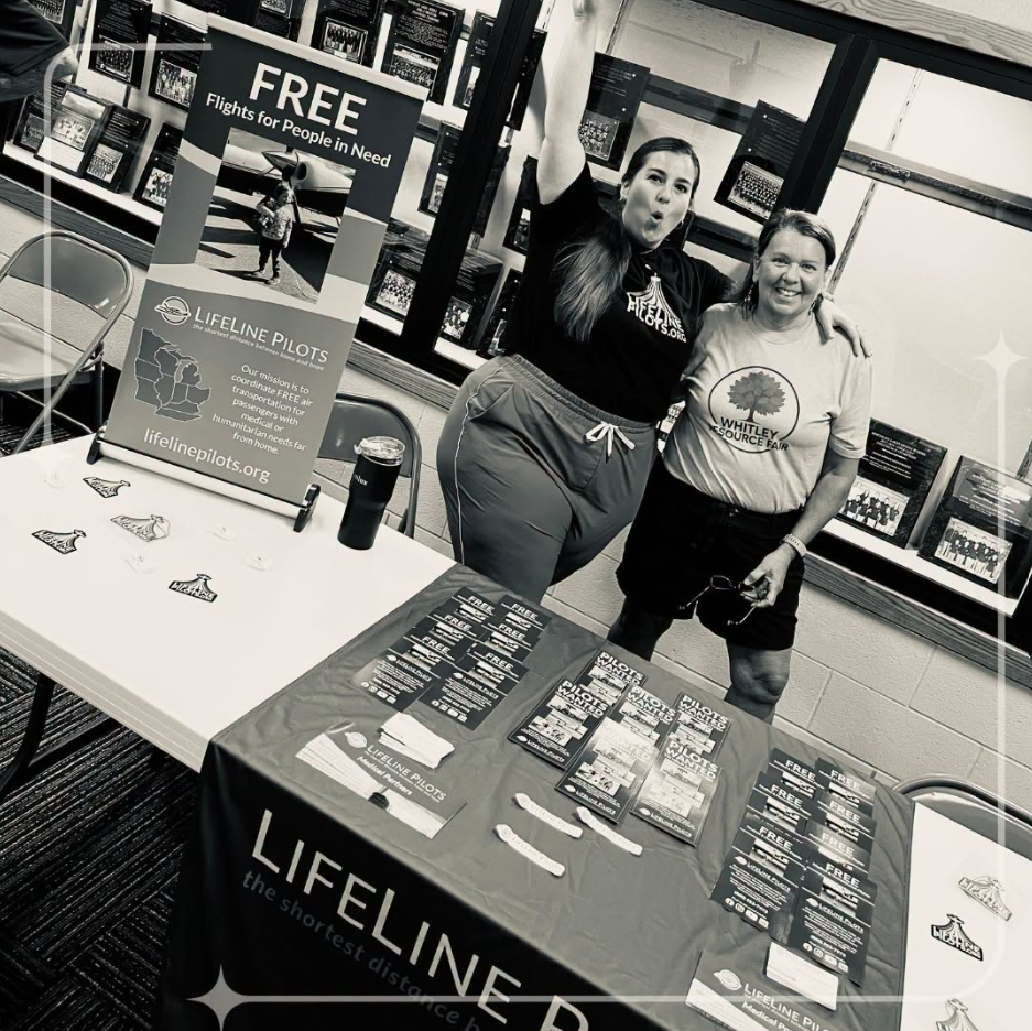 Black and white photograph of two women smiling behind a vendor table neatly laid out with a Lifeline banner and giveaway items
