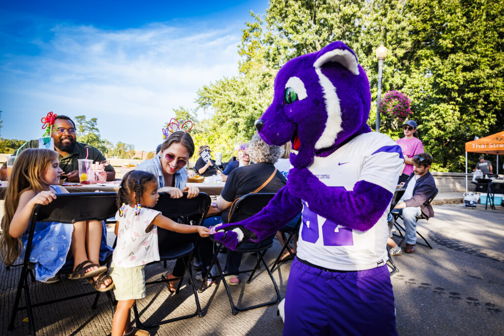 A purple cat mascot from Muncie Community Schools is handed something by a small child while adults watch on from a long table set up on the Washington Street Bridge